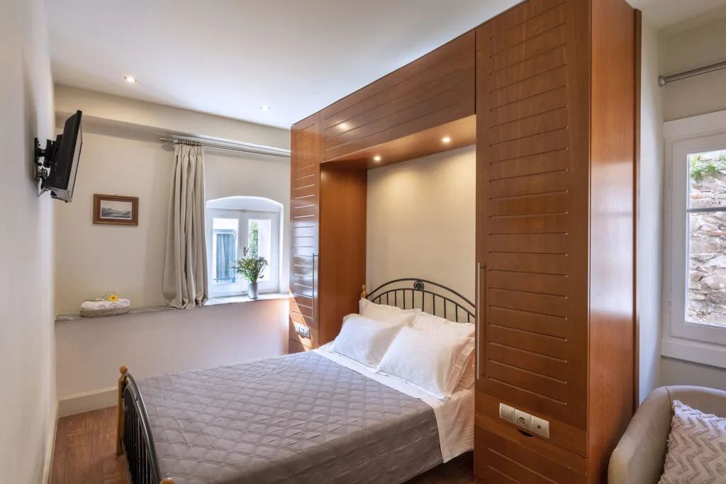 An interior view of a modern single hotel room at Hotel Palladion in Ermoupoli, Syros, featuring a comfortable bed with white linen and a grey throw, flanked by large wooden built-in cupboards and illuminated by warm recessed lighting.