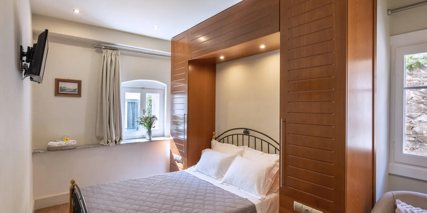 An interior view of a modern single hotel room at Hotel Palladion in Ermoupoli, Syros, featuring a comfortable bed with white linen and a grey throw, flanked by large wooden built-in cupboards and illuminated by warm recessed lighting.